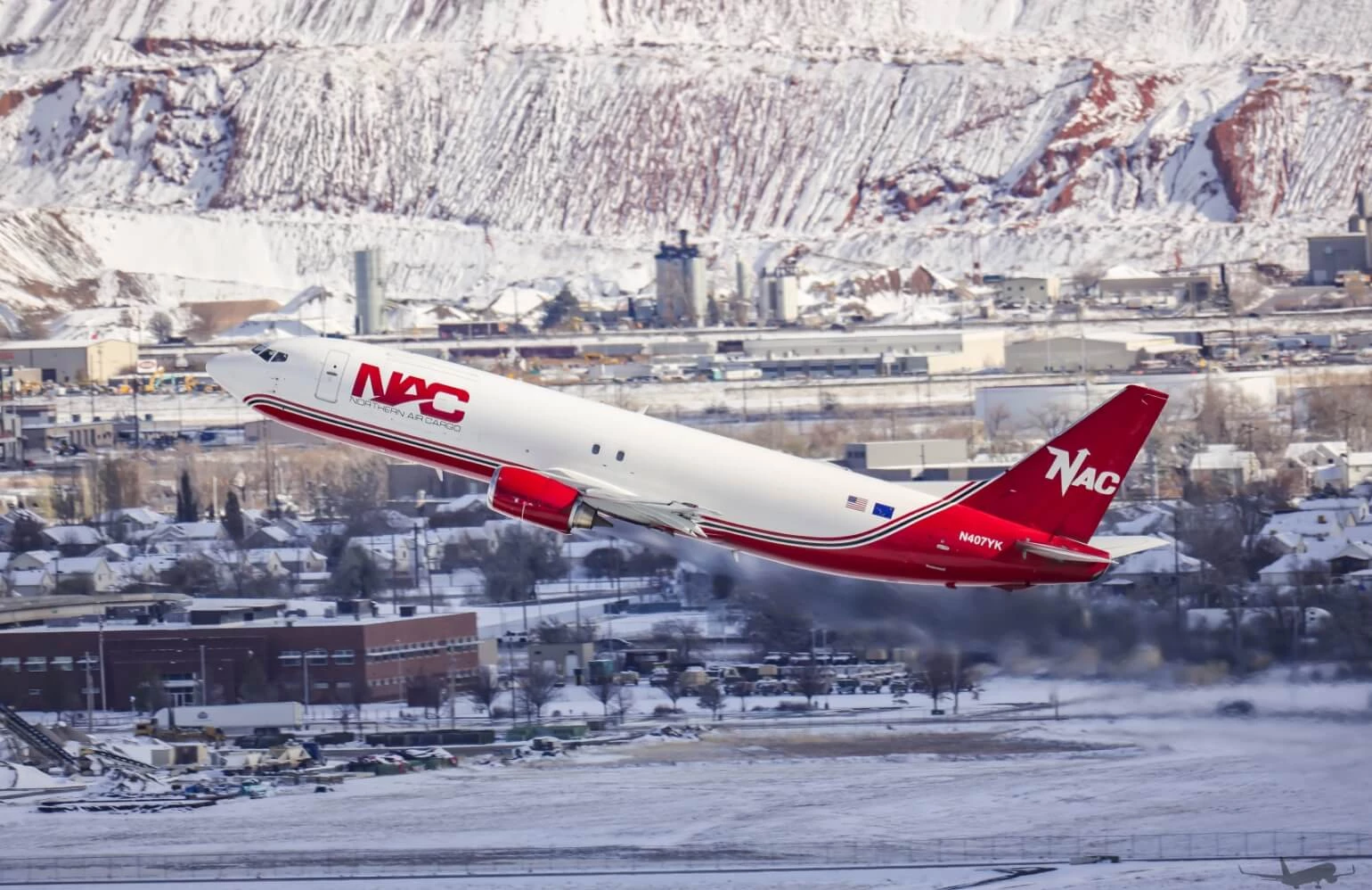 A Northern Air Cargo plane in flight with mountains and a city in the background.