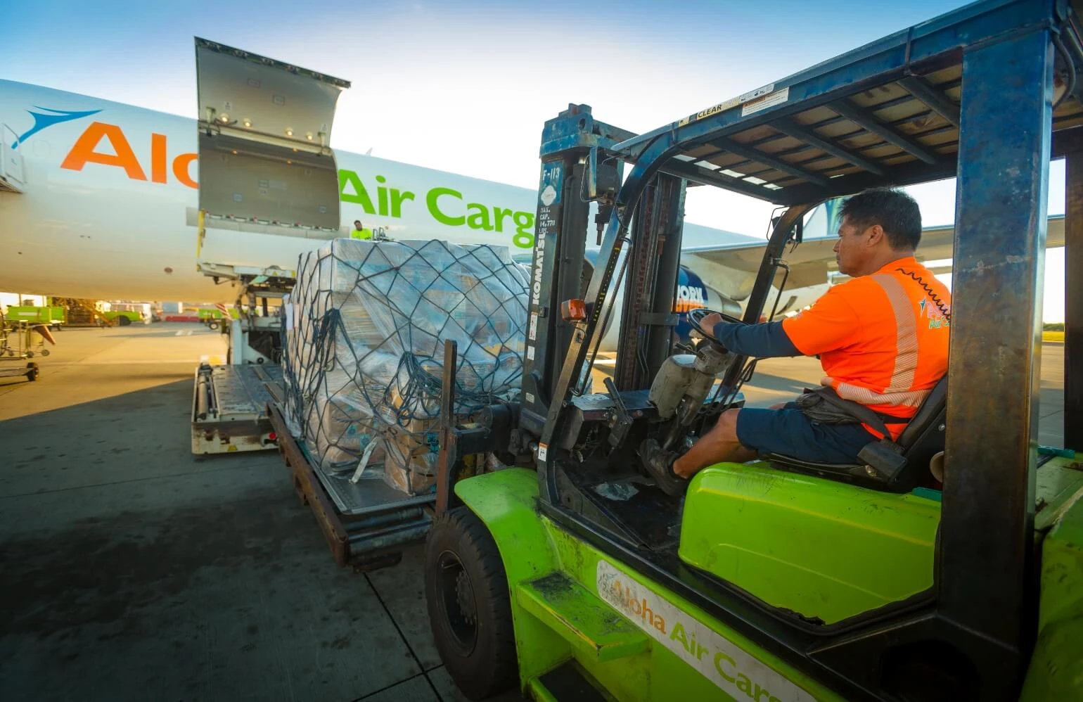 An Aloha Air Cargo customer service employee driving a forklift that is delivering cargo to a plane.