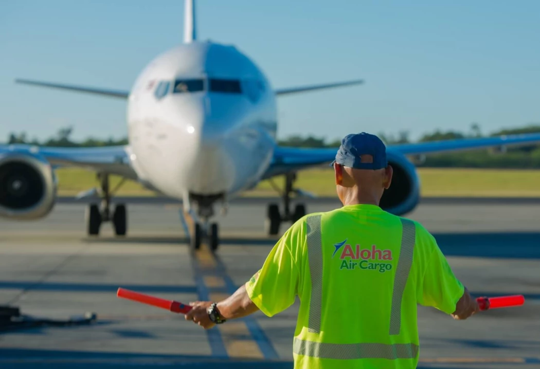 An Aloha Air Cargo employee using orange safety batons to help signal a plane on the runway.