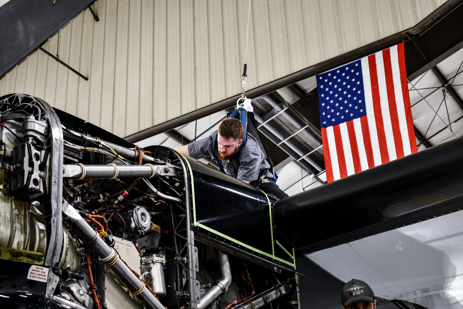 A Northern Air Maintenance Services employee using a safety harness to work on the top of a plane. An American flag hangs in the background.