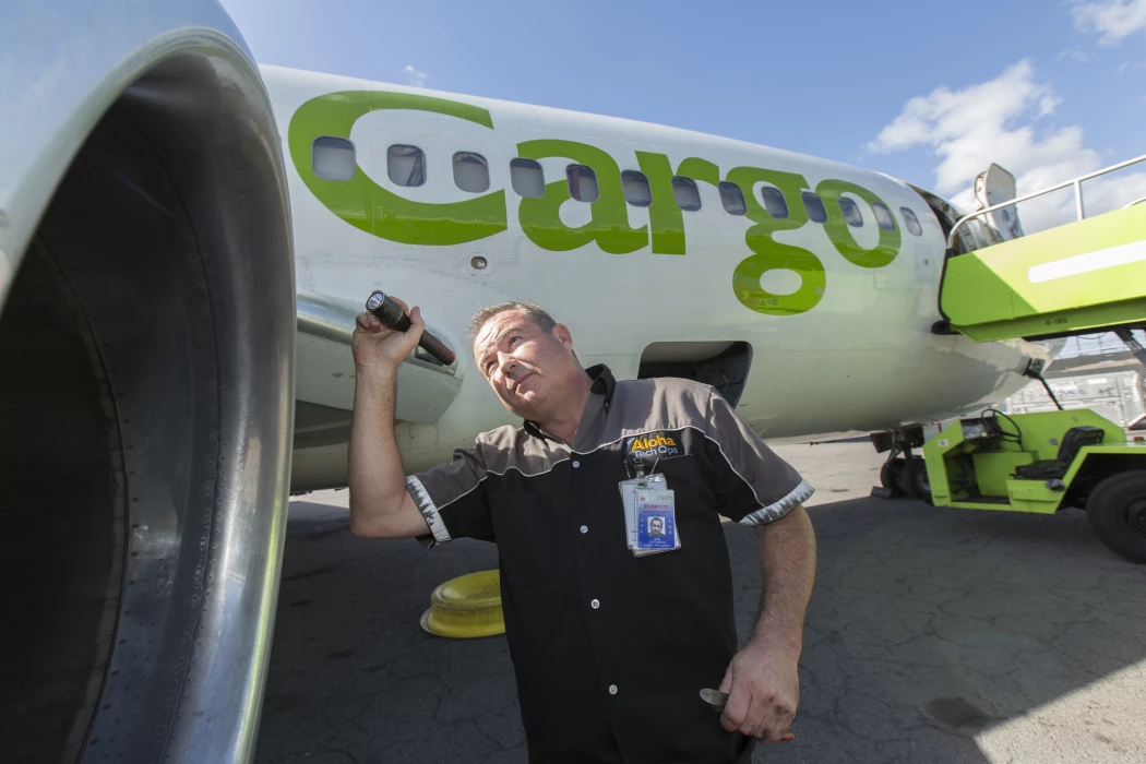 An Aloha Tech Ops employee uses a flashlight to look at a plane's engine.